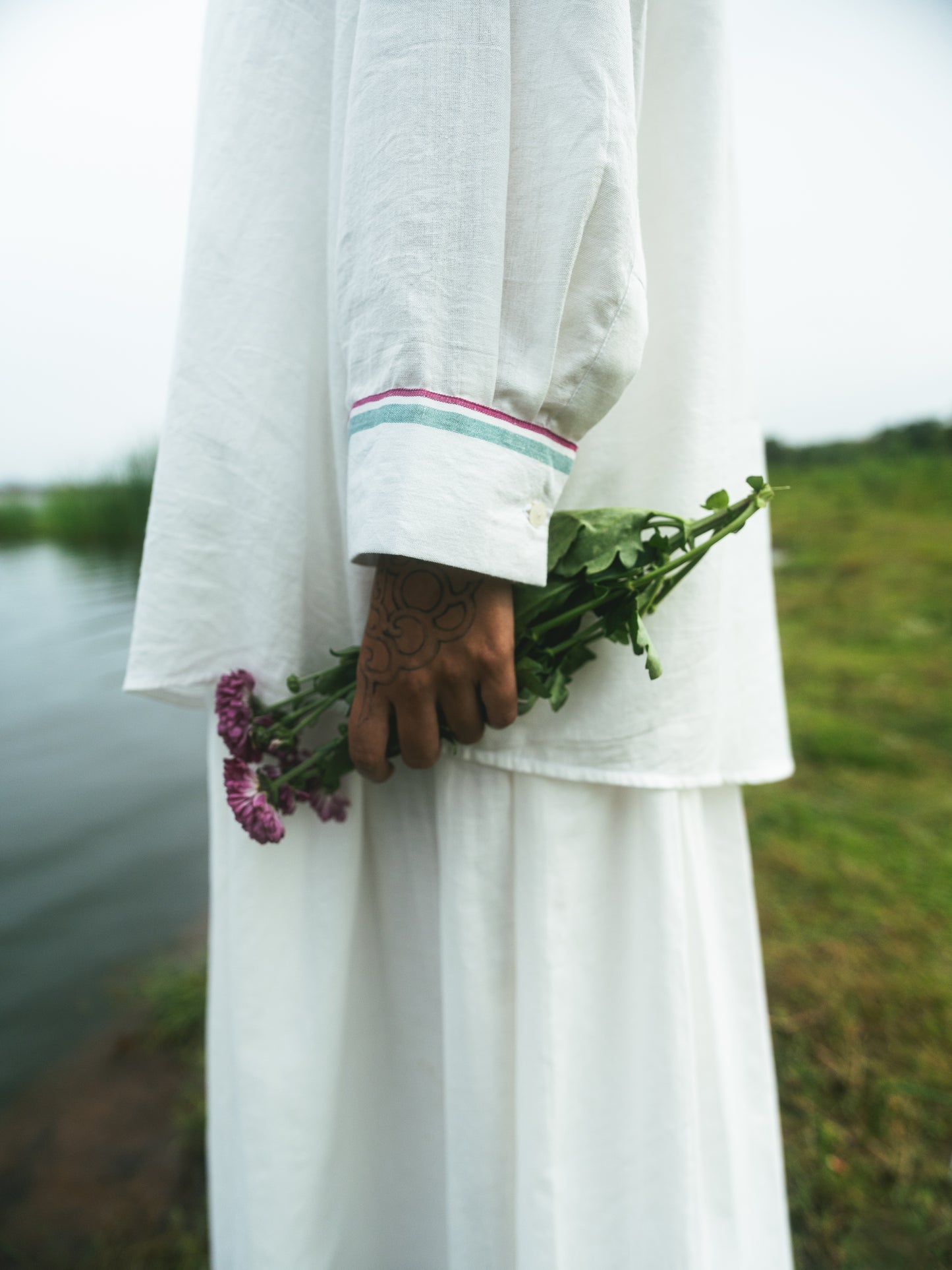 Person in a white outfit holding a bundle of flowers by a body of water.