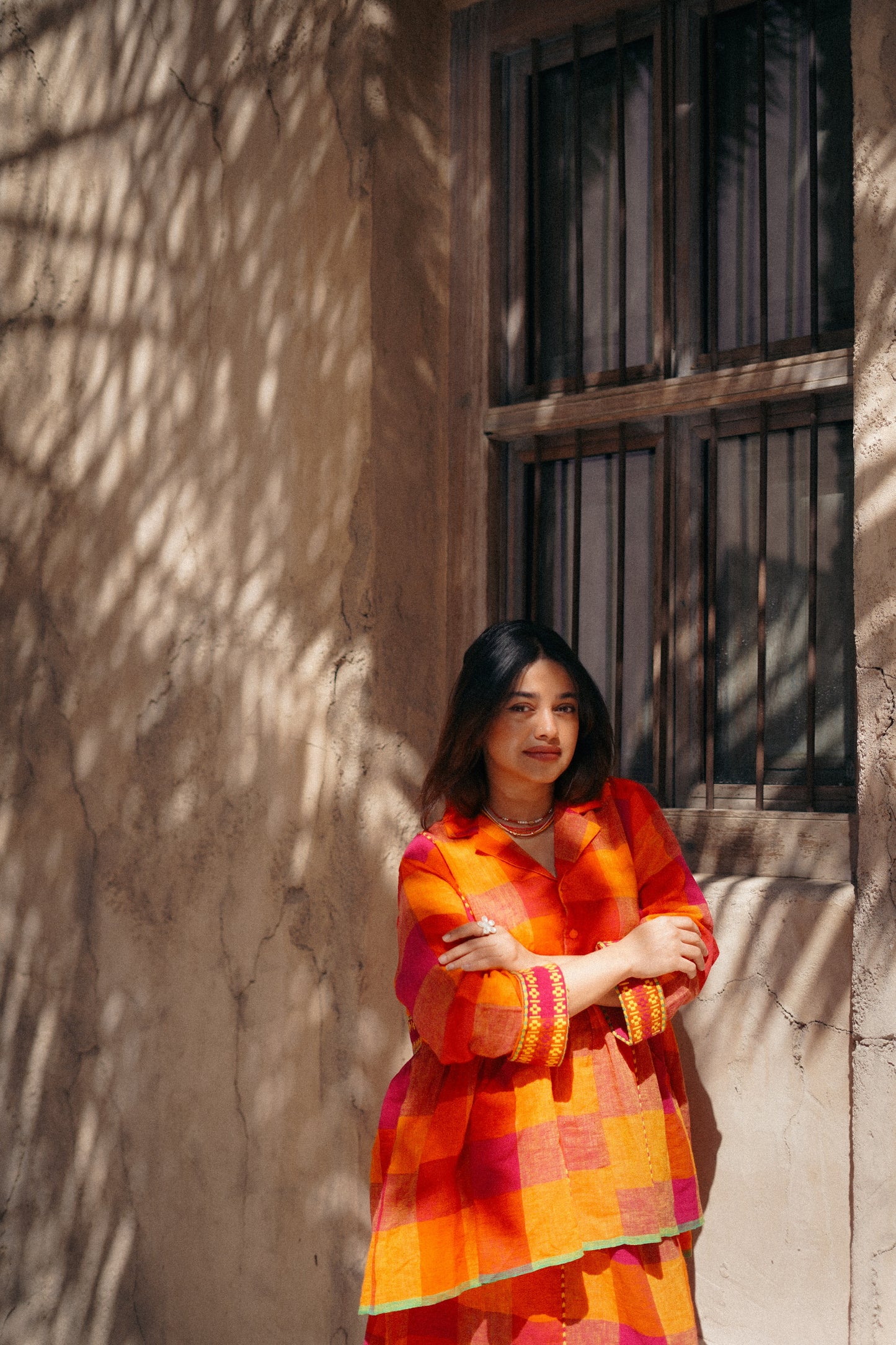 Woman in a colorful dress standing against a textured wall with a window.