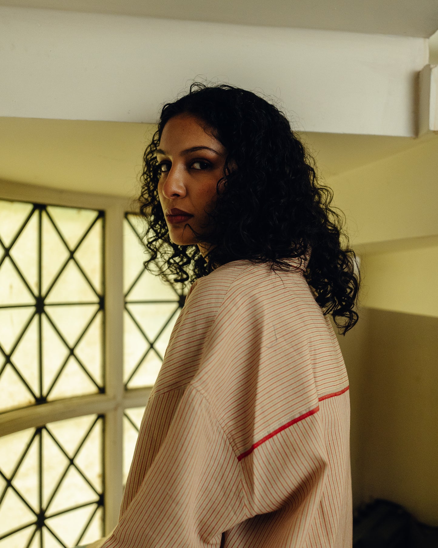 Woman in a beige Oversized Shirt - Sand Striped with red trim standing in a room with a window in the background