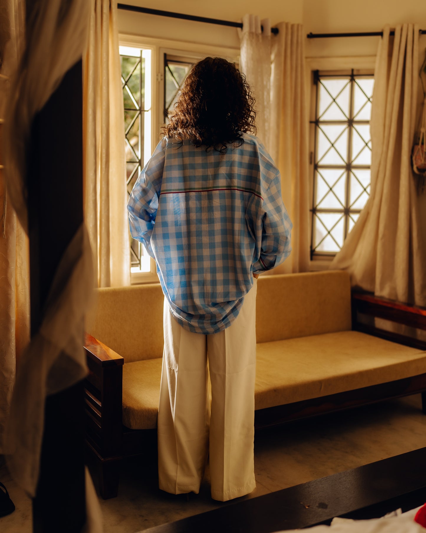 Person wearing a Oversized shirt - Light blue checks and white pants standing in a room with curtains and a sofa.