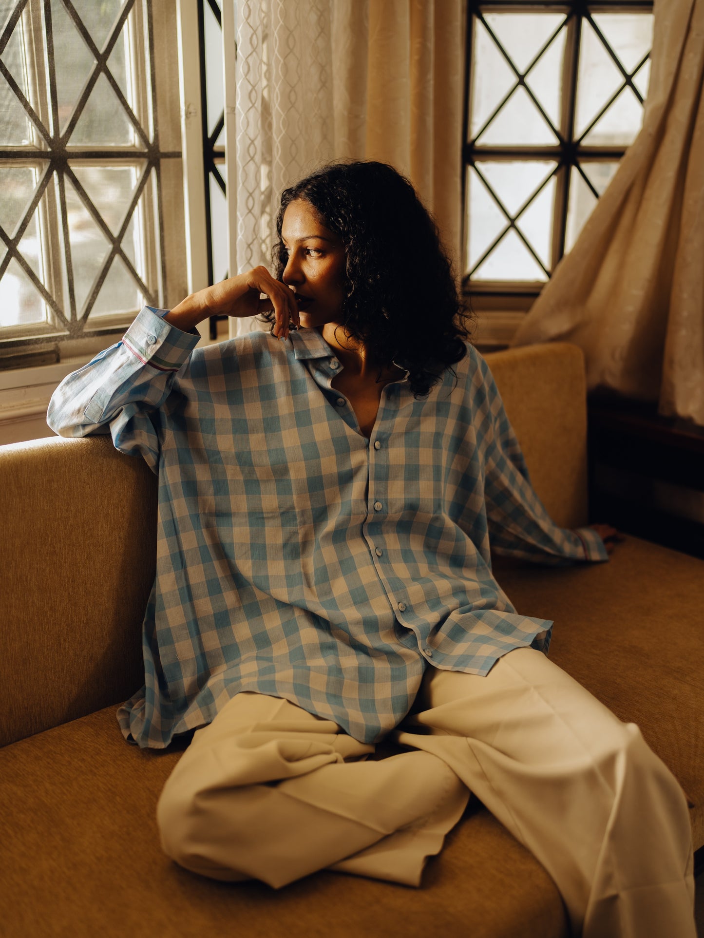 Woman sitting on a couch wearing a Oversized shirt - Light blue checks and beige pants, with a window in the background.