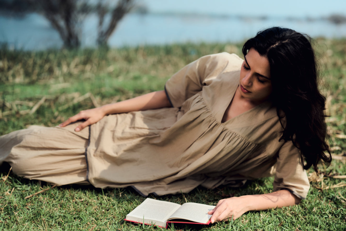 Woman in a beige dress lying on grass reading a book with a natural background