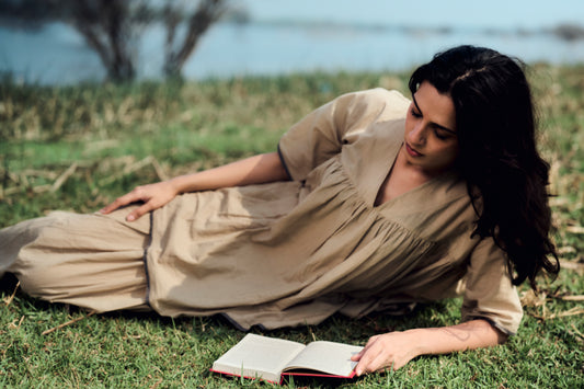 Woman in a beige dress lying on grass reading a book with a natural background