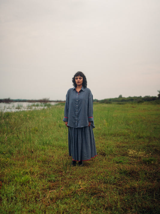 Person wearing a blue checkered outfit standing in a grassy field with water and sky in the background