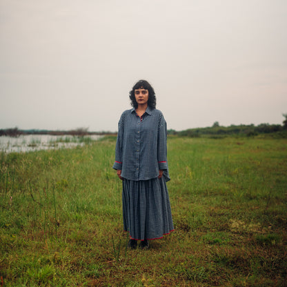 Person wearing a checkered outfit standing in a grassy field with a body of water and trees in the background.