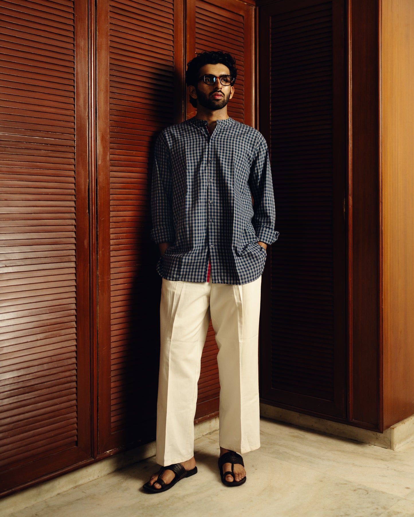 Man wearing a Blueberry checkered full sleeve shirt and white pants standing against a wooden door.