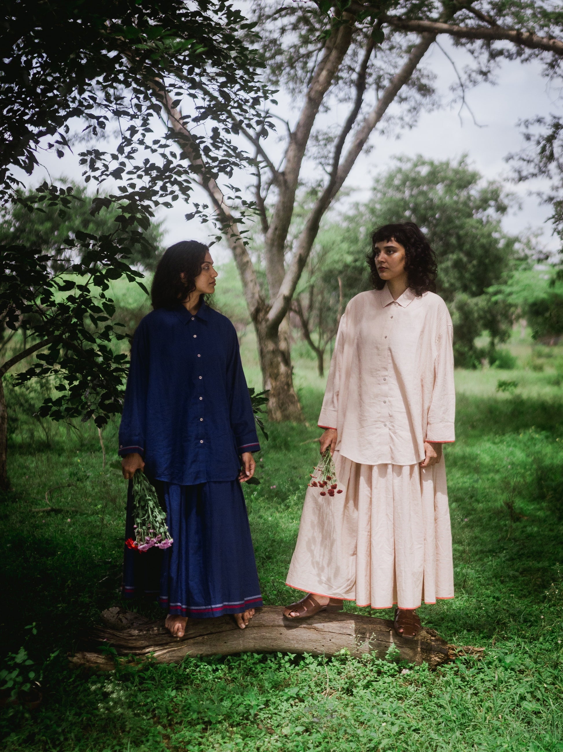 Two women standing in a forested area, one in a blue outfit and the other in a beige outfit.