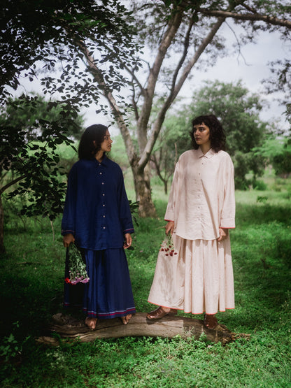 Two women standing in a forested area, one in a blue outfit and the other in a beige outfit.