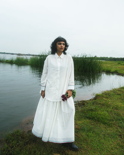 Person wearing a white outfit standing by a body of water with greenery in the background