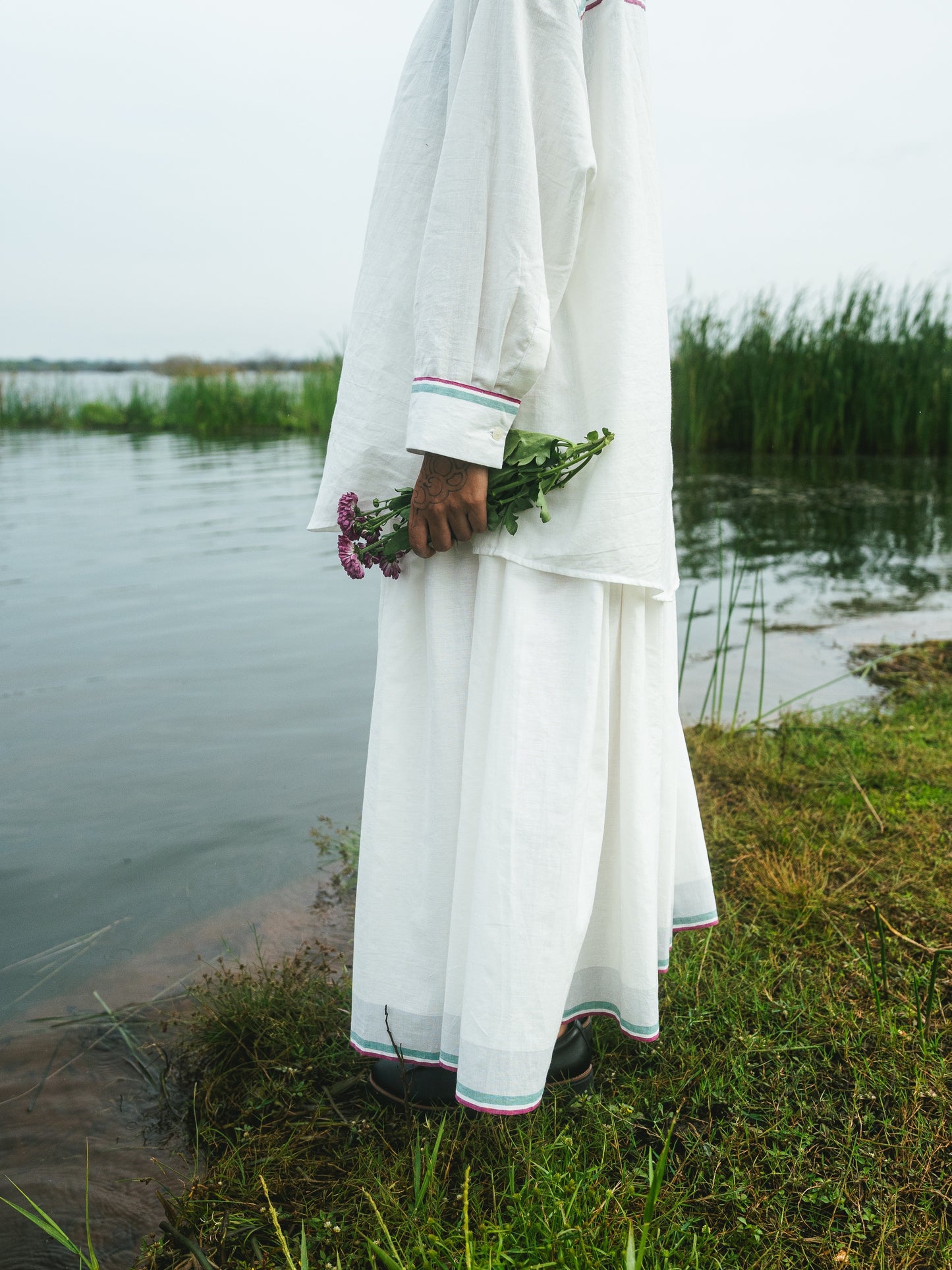 Person in a white traditional outfit holding flowers by a body of water