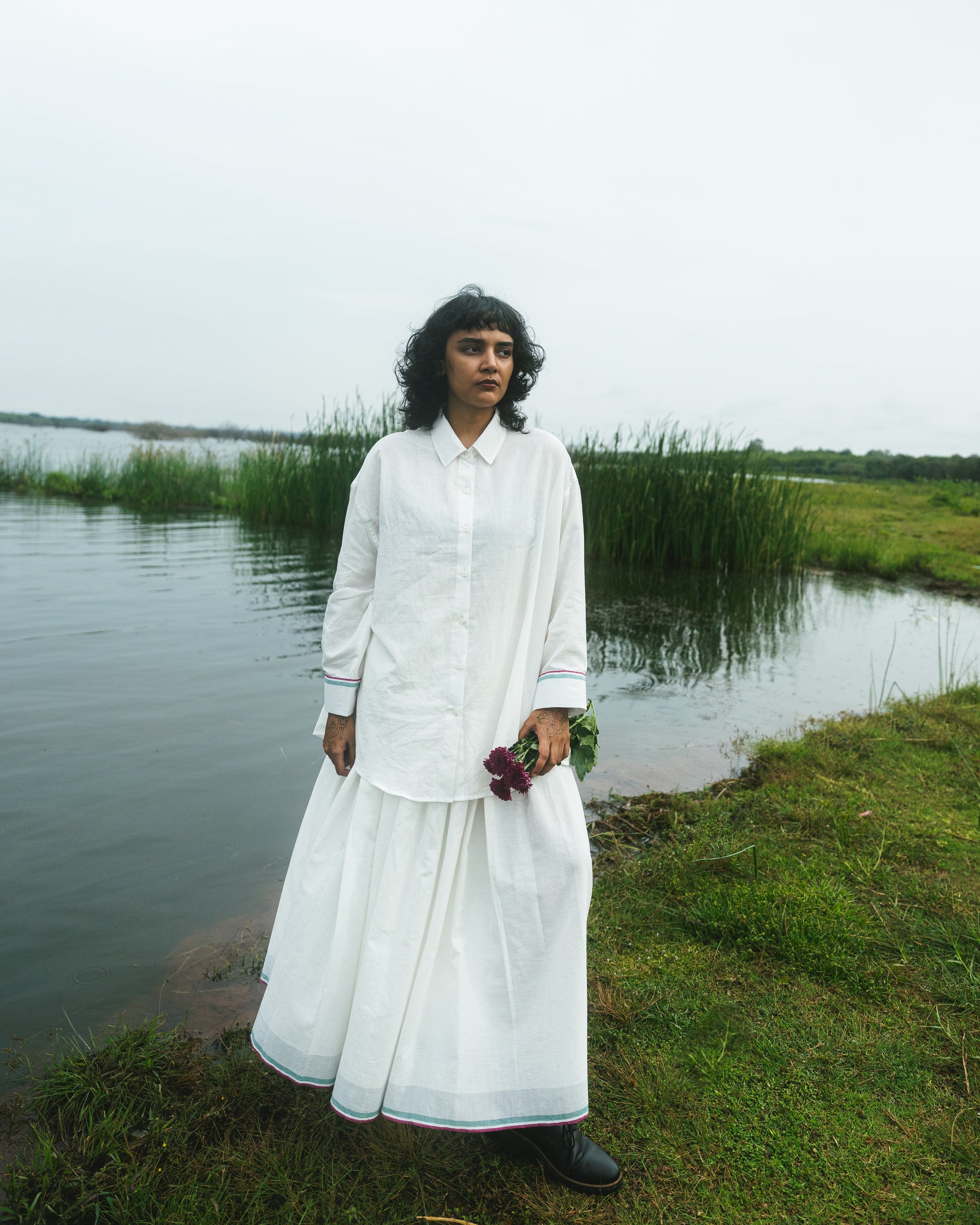 Person in a white outfit standing by a body of water with greenery in the background