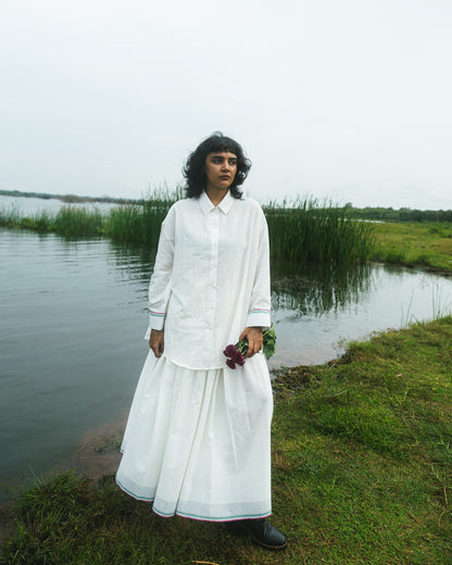 Person in a white outfit standing by a body of water with greenery in the background