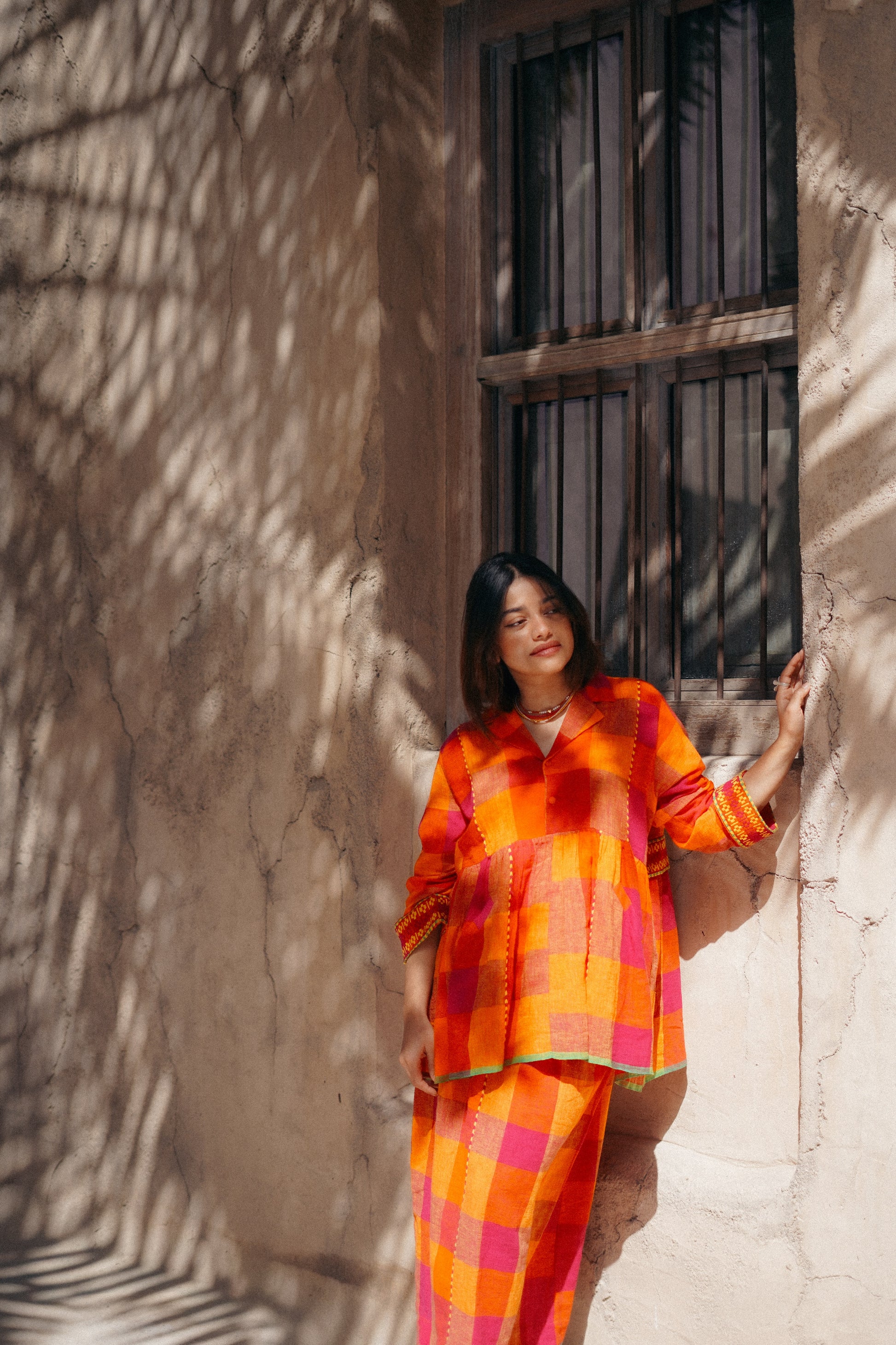 Woman in a bright orange and pink checkered outfit standing against a textured wall with a window.