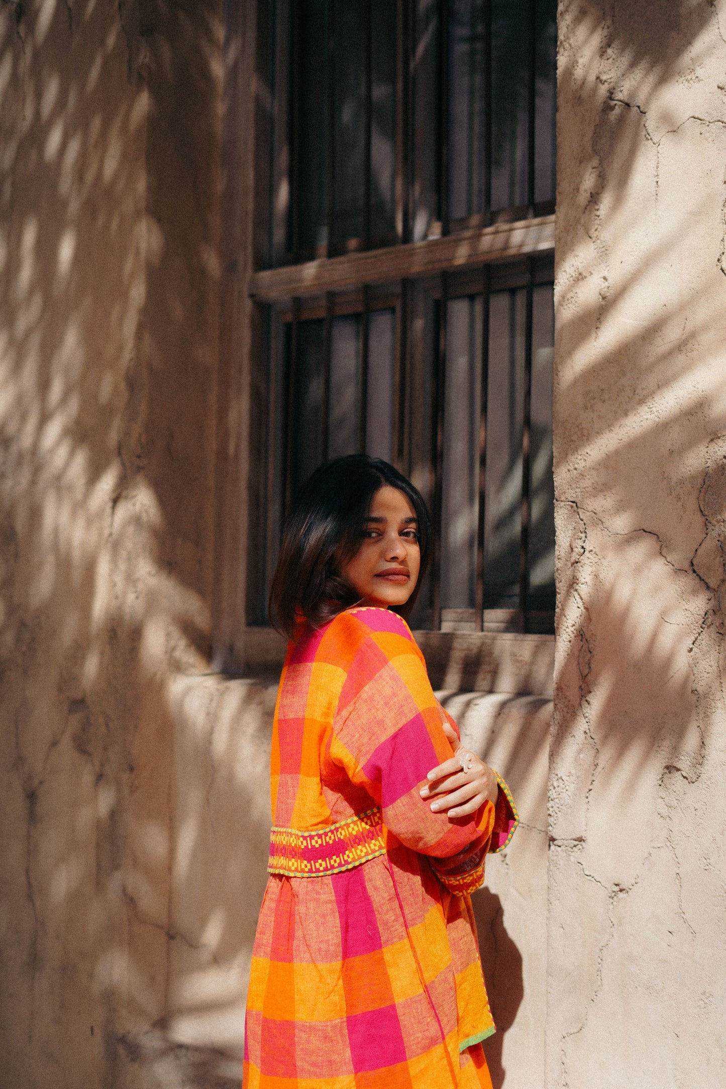 Woman in a colorful dress standing against a textured wall with a window.