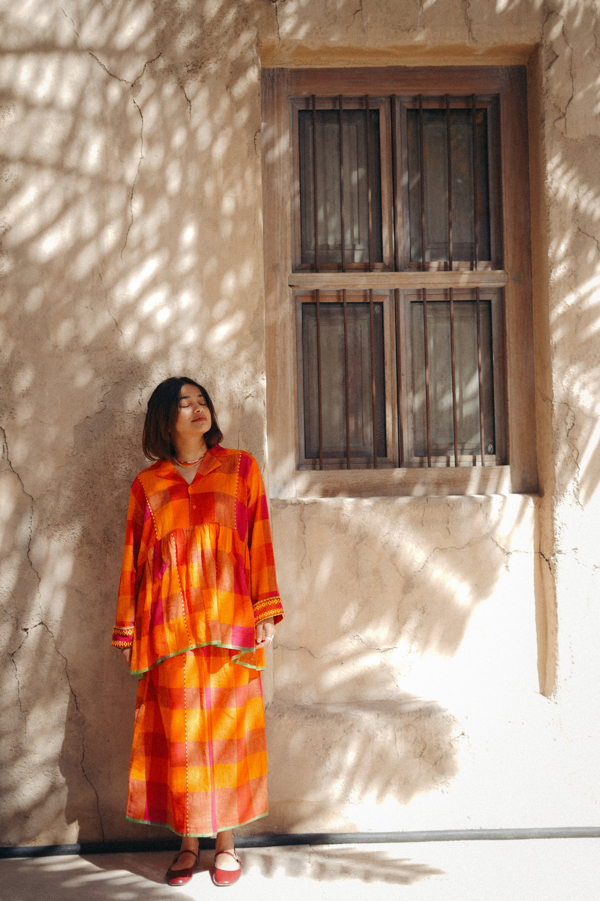 Woman in an orange dress standing in front of a textured wall with a window.