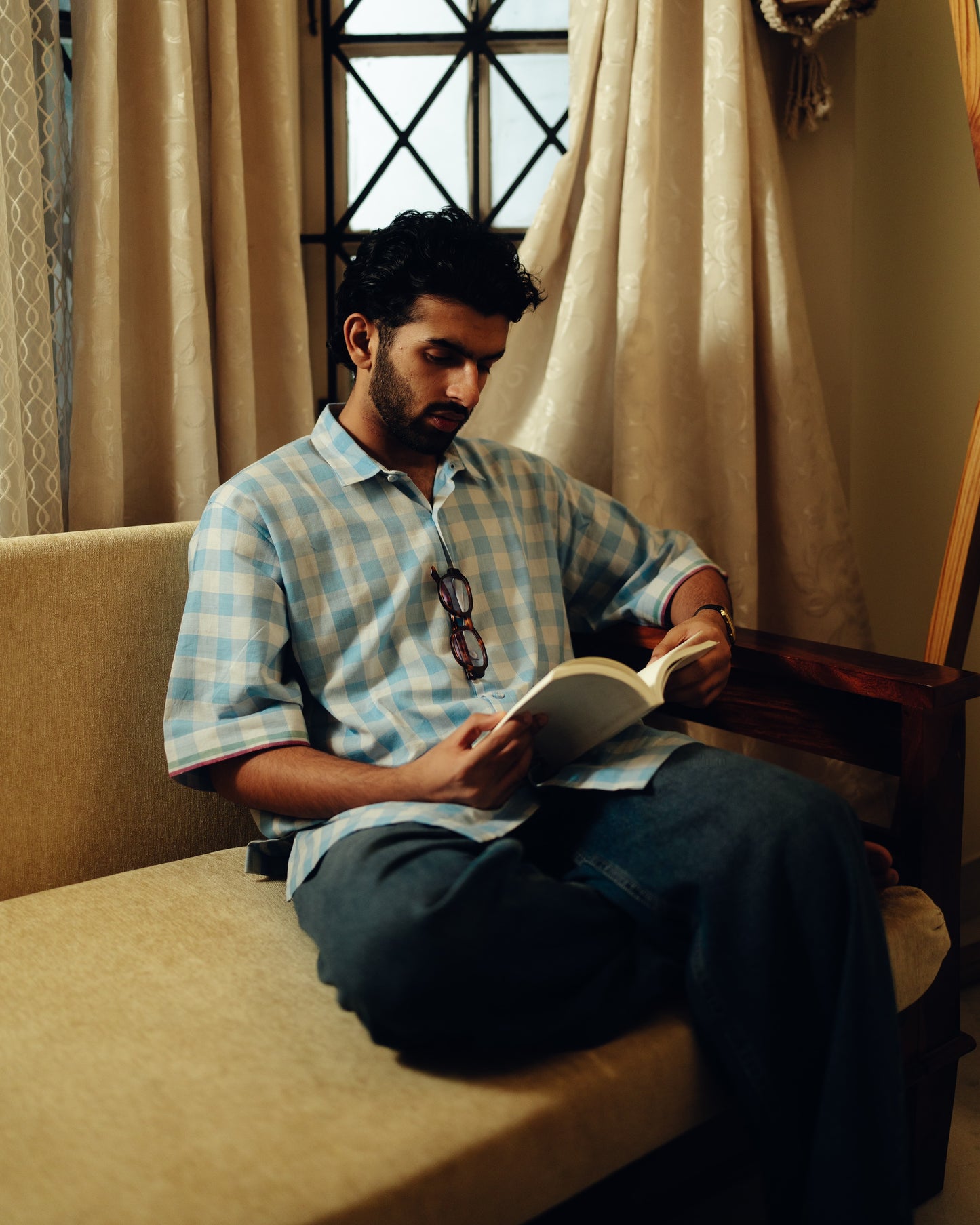 Man reading a book on a couch with curtains and a window in the background