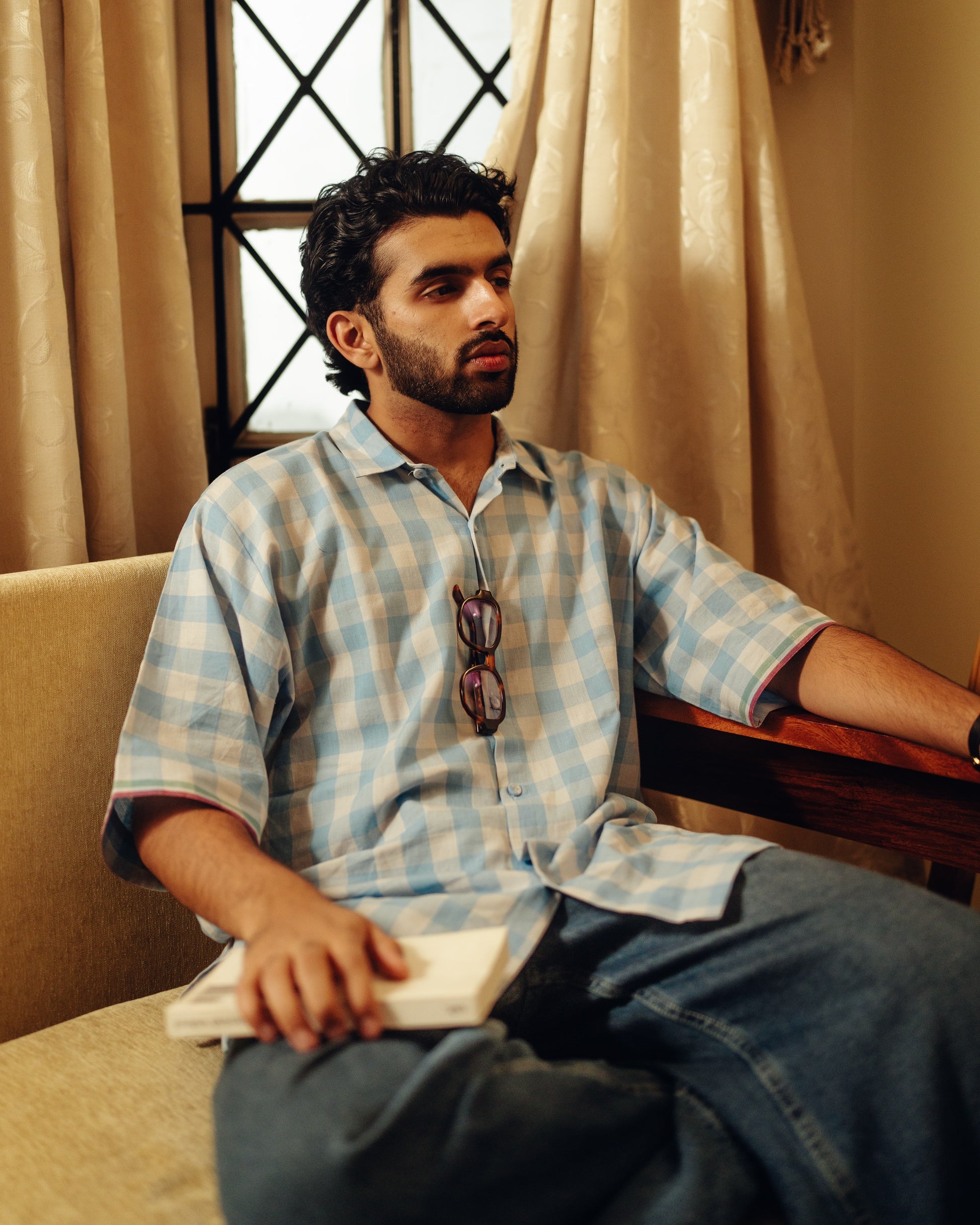 Man sitting on a couch wearing a checkered Half-sleeves shirt - Light blue checks with sunglasses on his shirt, in a room with beige curtains.