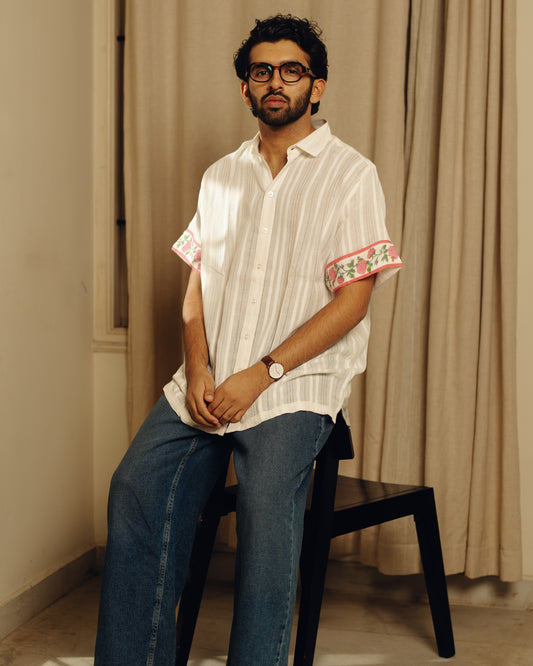 Man sitting on a chair wearing a white striped shirt with floral cuffs and blue jeans.