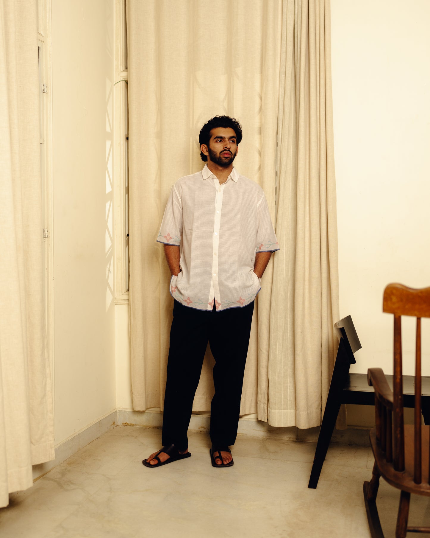 Man standing in a room with light-colored curtains and a wooden chair.