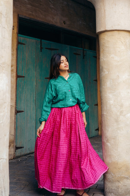 Woman wearing a green top and pink skirt standing in front of a stone wall and wooden door.