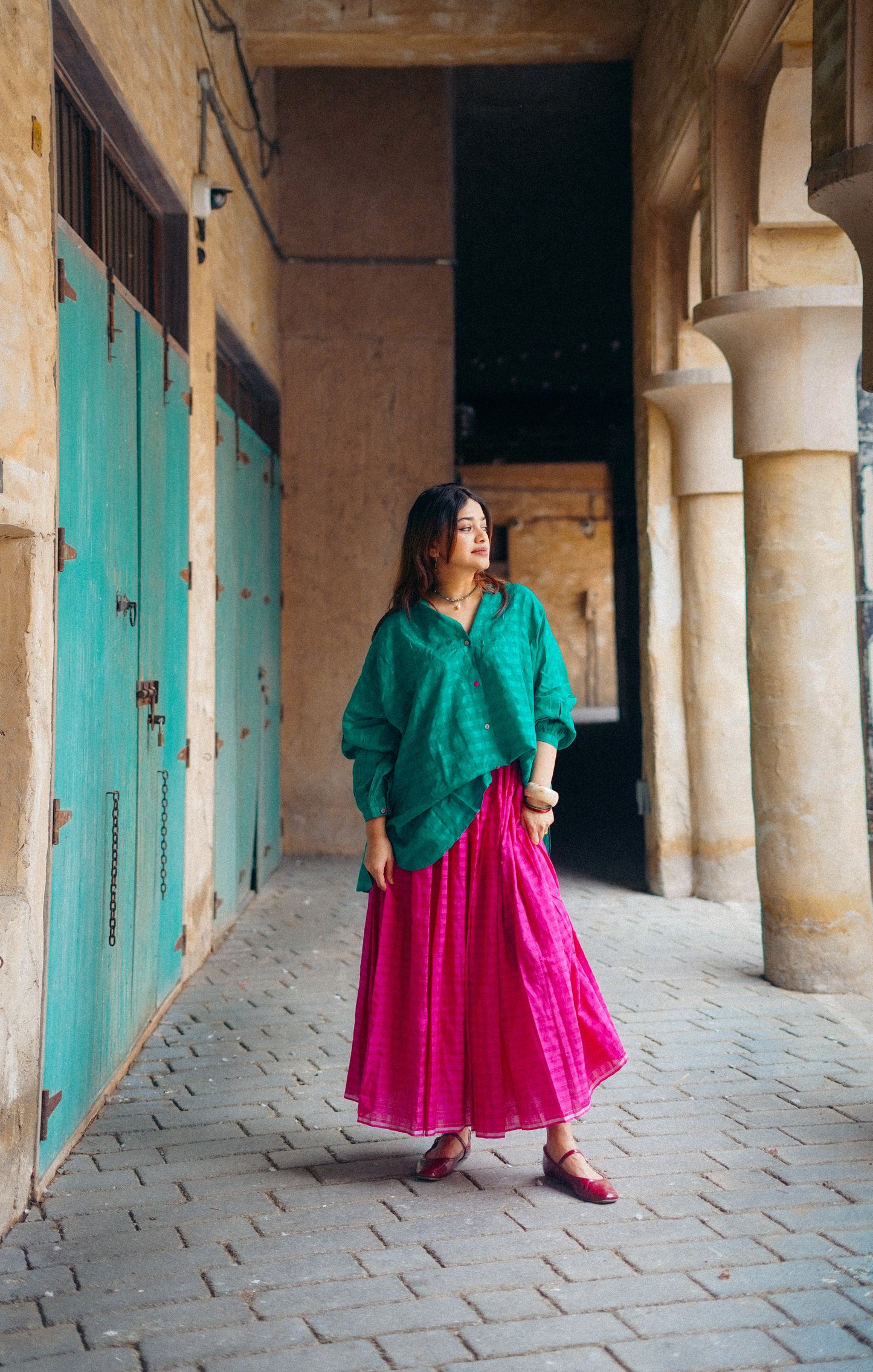 Woman in green top and pink skirt standing in a sunlit hallway with columns and turquoise doors.