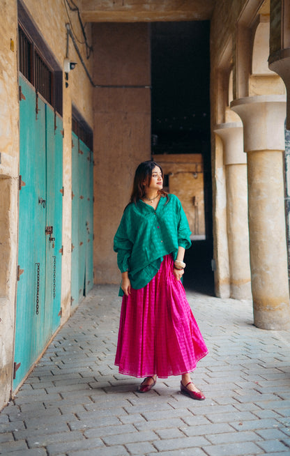 Woman in green top and pink skirt standing in a sunlit hallway with columns and turquoise doors.