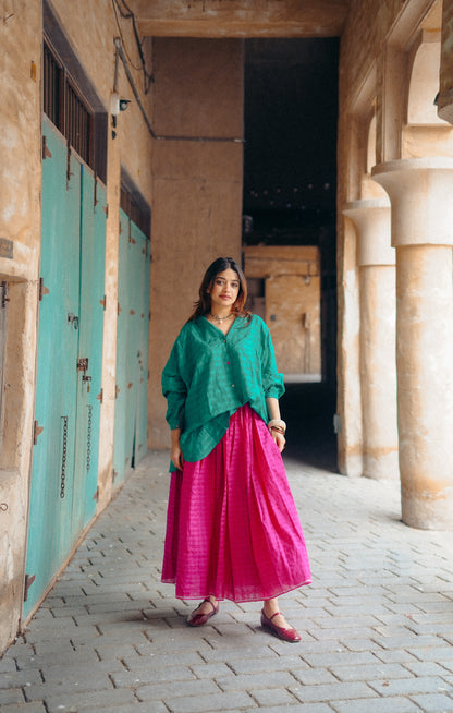 Woman in green top and pink skirt standing in a sunlit outdoor setting with columns and a blue door.