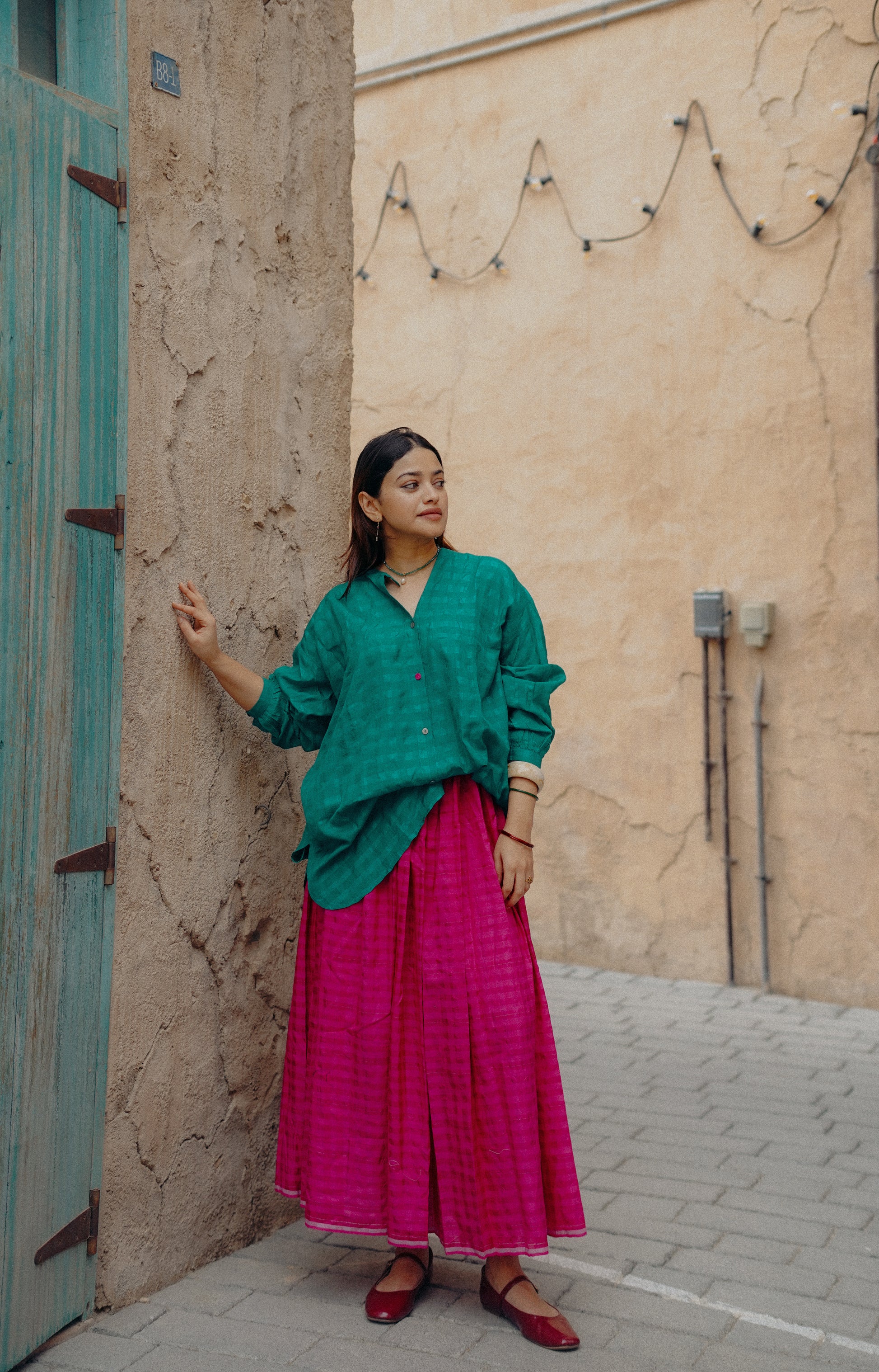 Woman in green top and pink skirt standing against a textured wall.