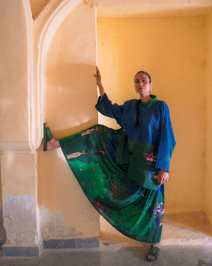 Woman in a blue and green traditional outfit standing in a yellow-walled room.