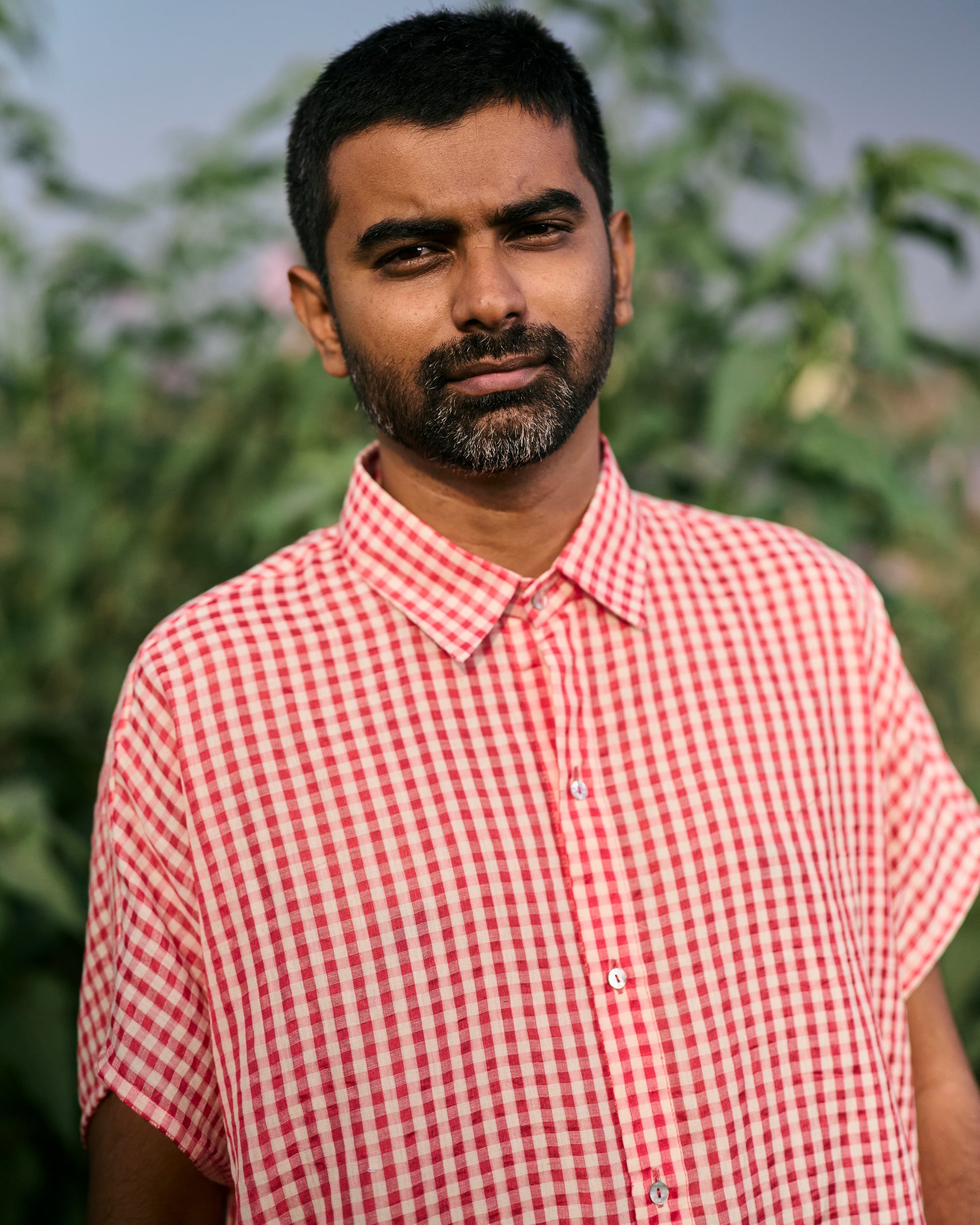 Man wearing a red and white checkered shirt outdoors with greenery in the background