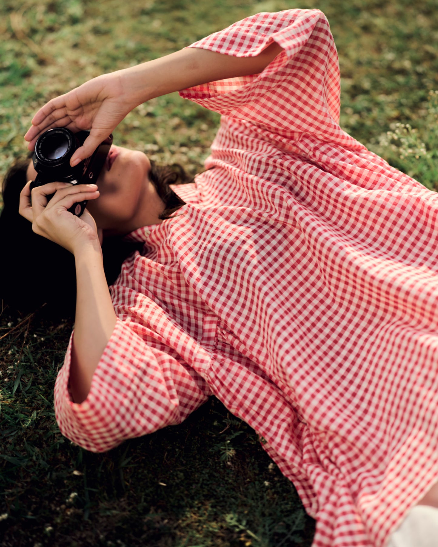 Person in a red and white checkered dress lying on grass, holding a camera.