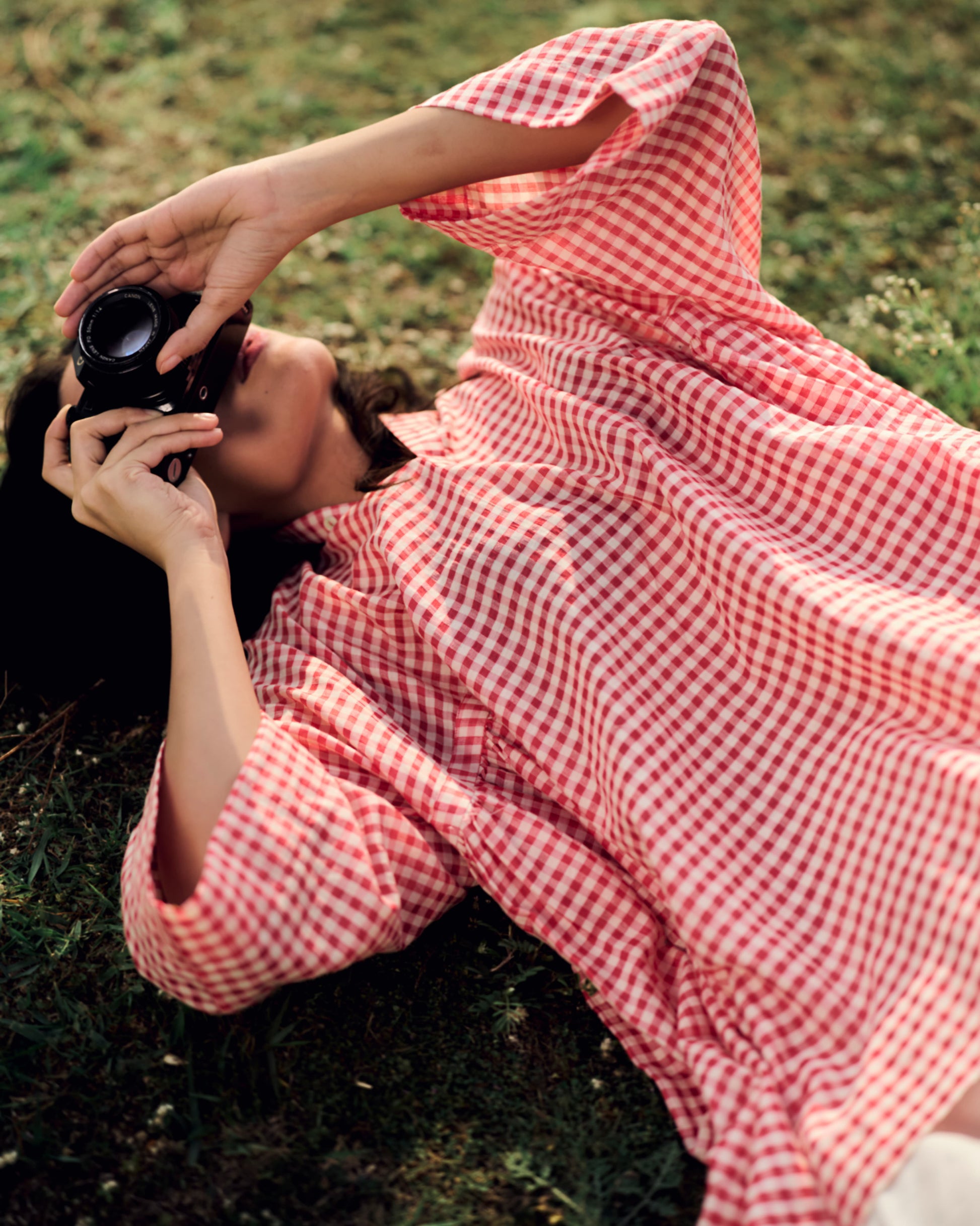 Person in a red and white checkered dress lying on grass, holding a camera.