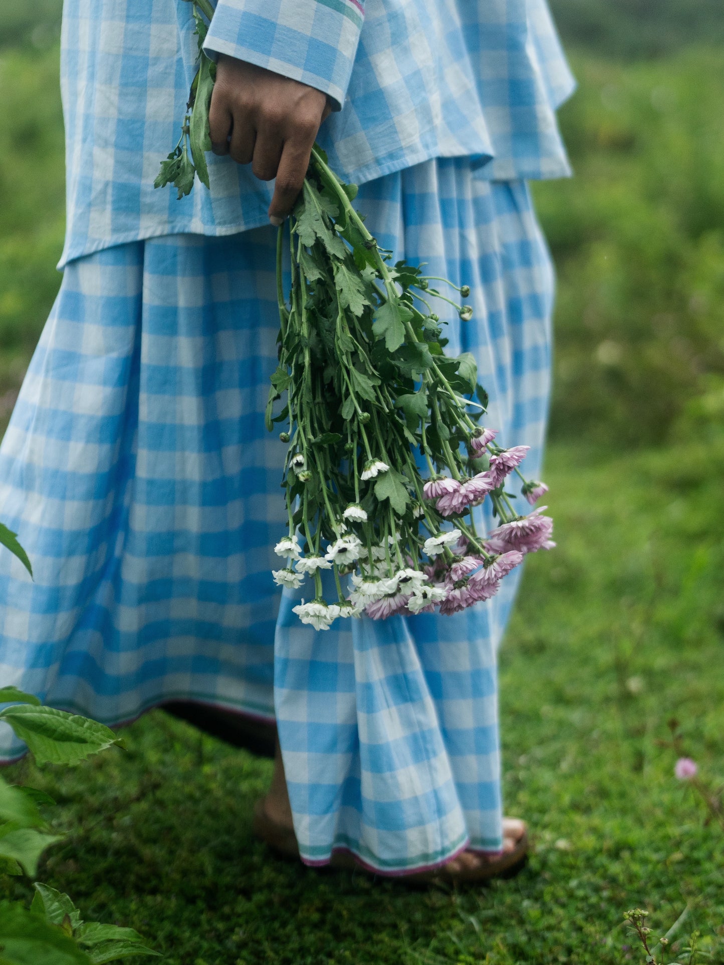 Person in a blue checkered outfit holding a bundle of flowers in a grassy field