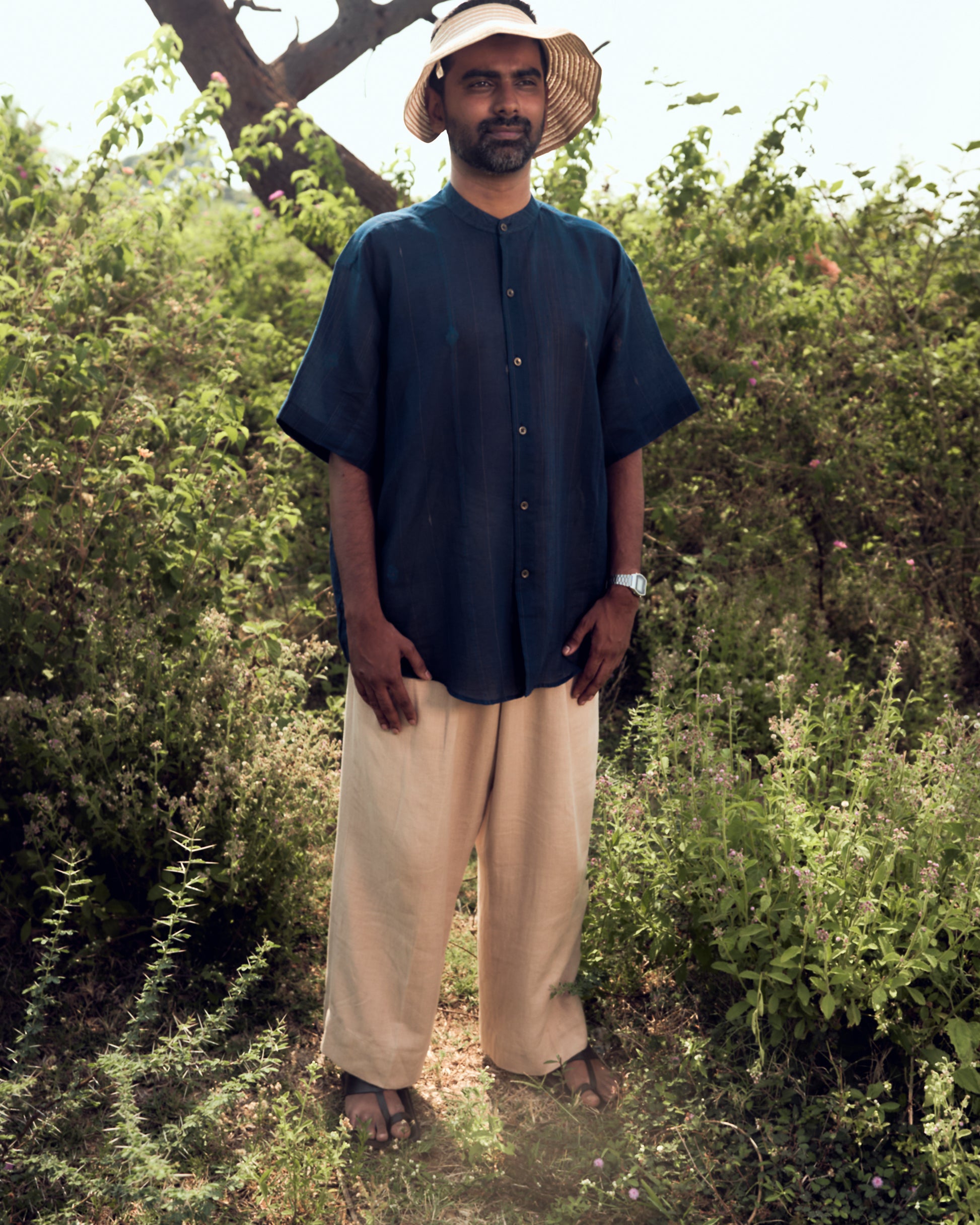 Man wearing a dark blue shirt and beige pants standing in a natural setting with greenery.