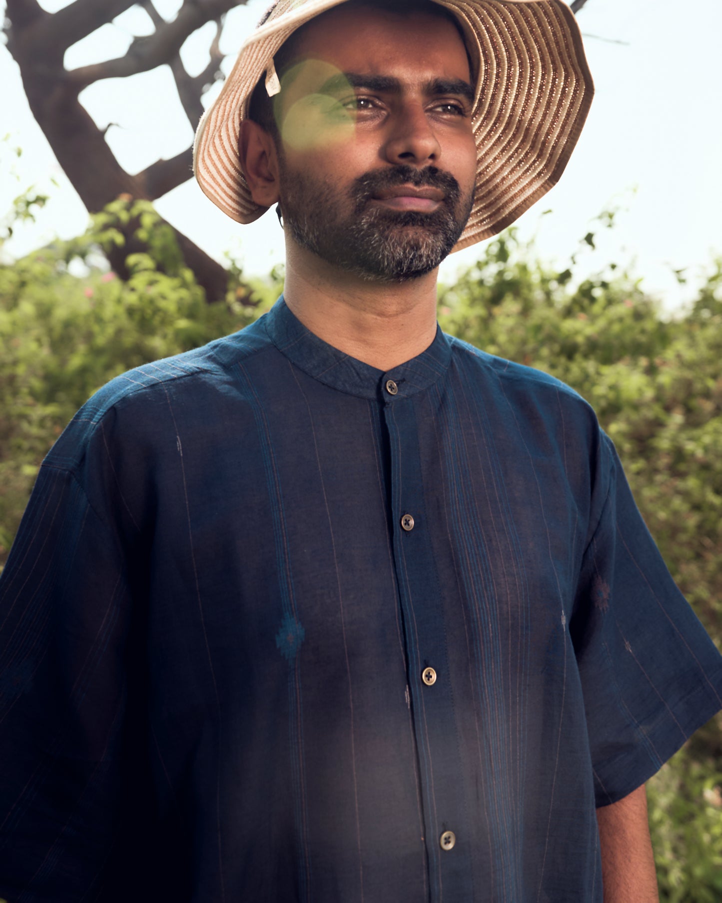 Man wearing a straw hat and dark blue shirt outdoors with greenery in the background