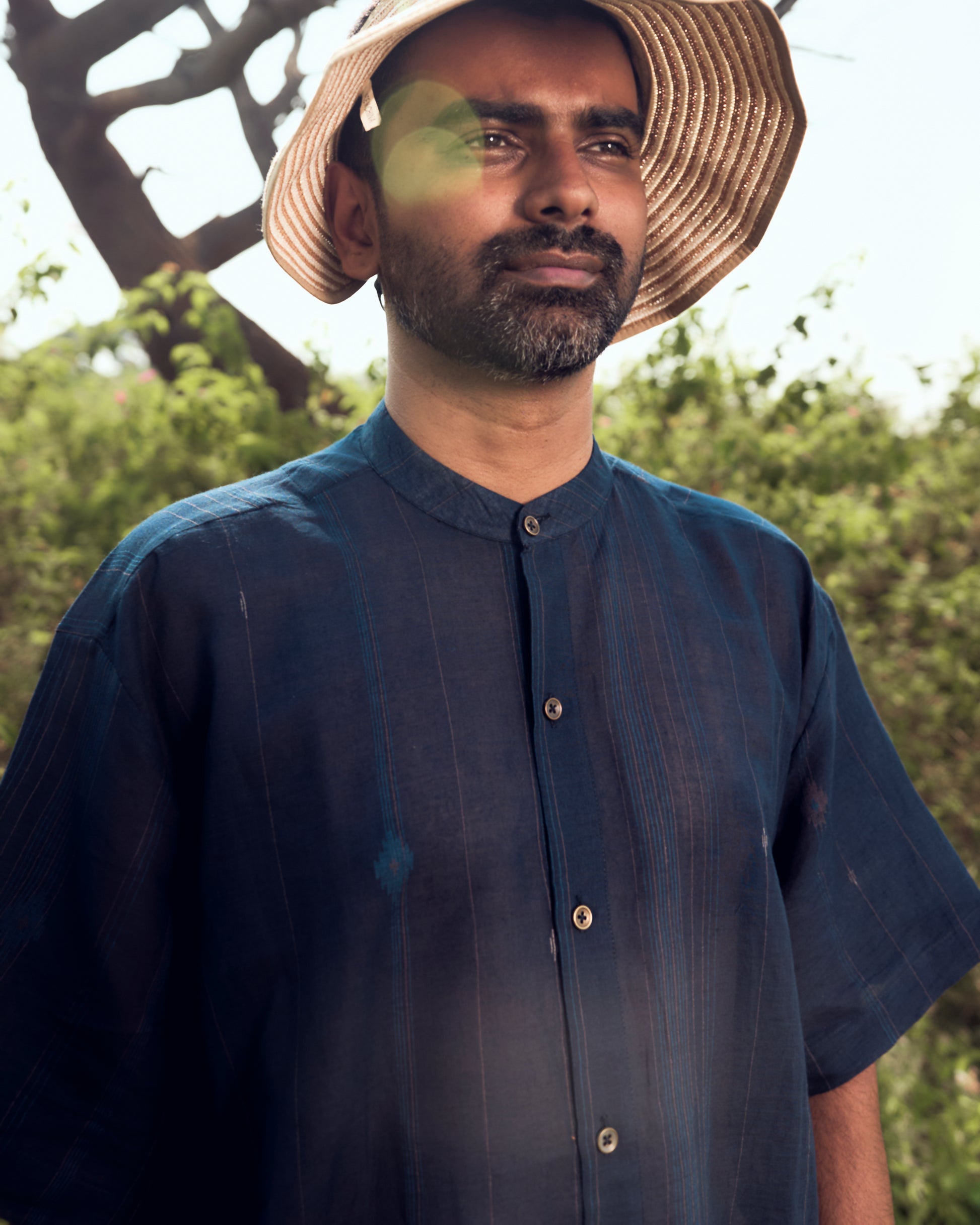 Man wearing a straw hat and dark blue shirt outdoors with greenery in the background