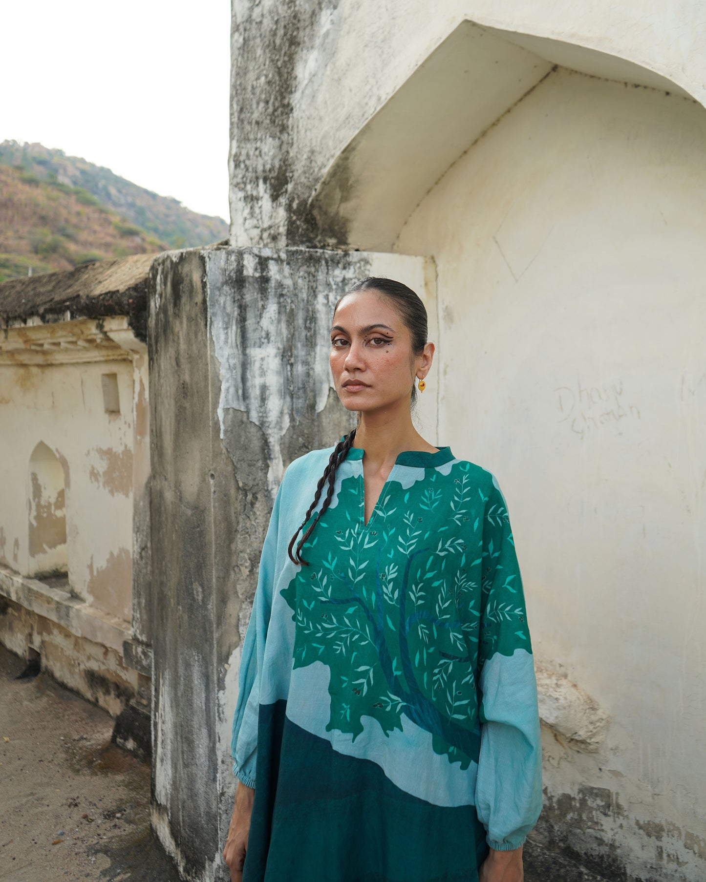 Woman wearing a green and blue patterned top standing against a weathered wall.