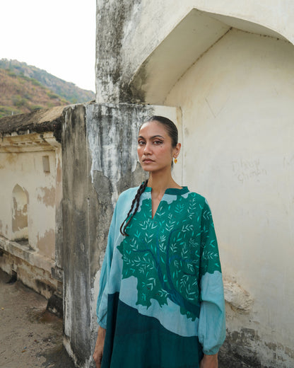 Woman wearing a green and blue patterned top standing against a weathered wall.