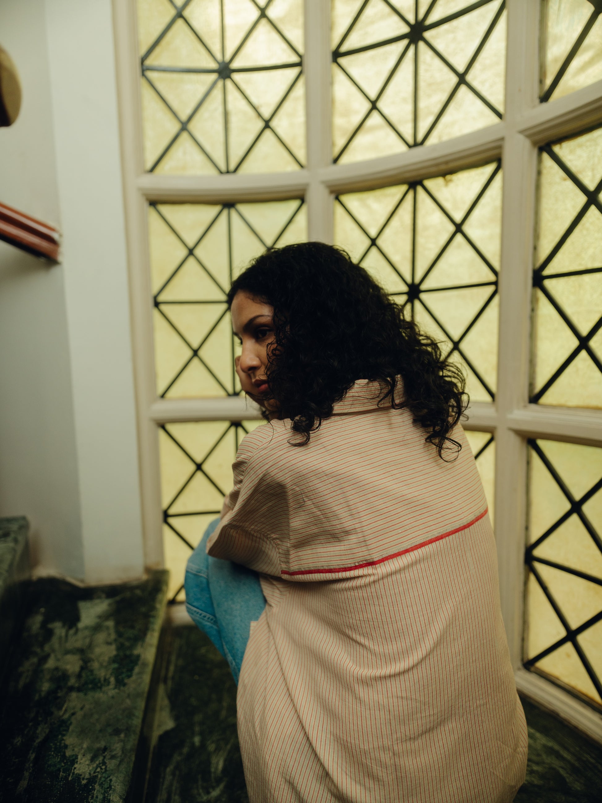 Woman wearing a Oversized Shirt - Sand Striped sitting in a room with geometric window design