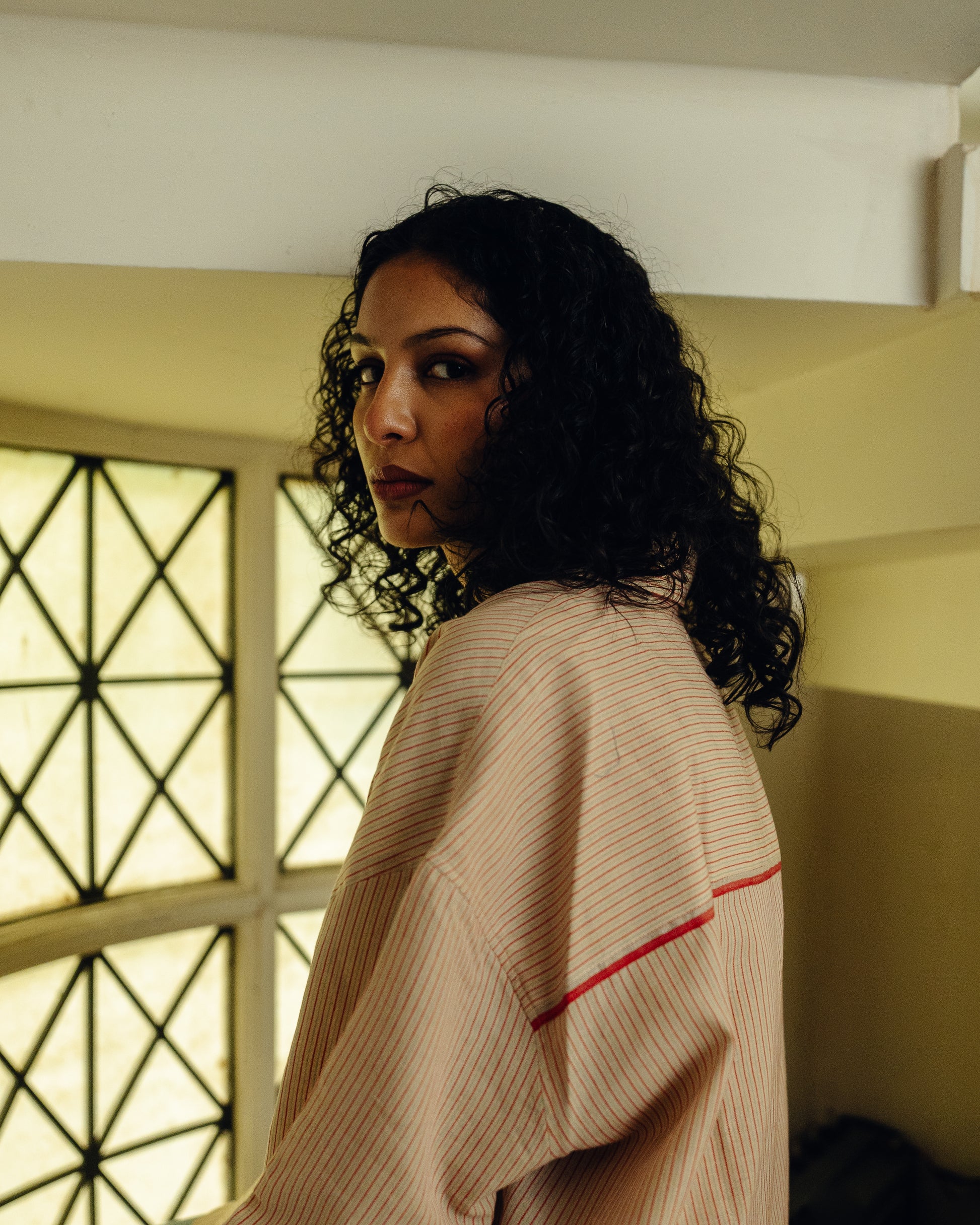Woman in a beige Oversized Shirt - Sand Striped with red trim standing in a room with a window in the background