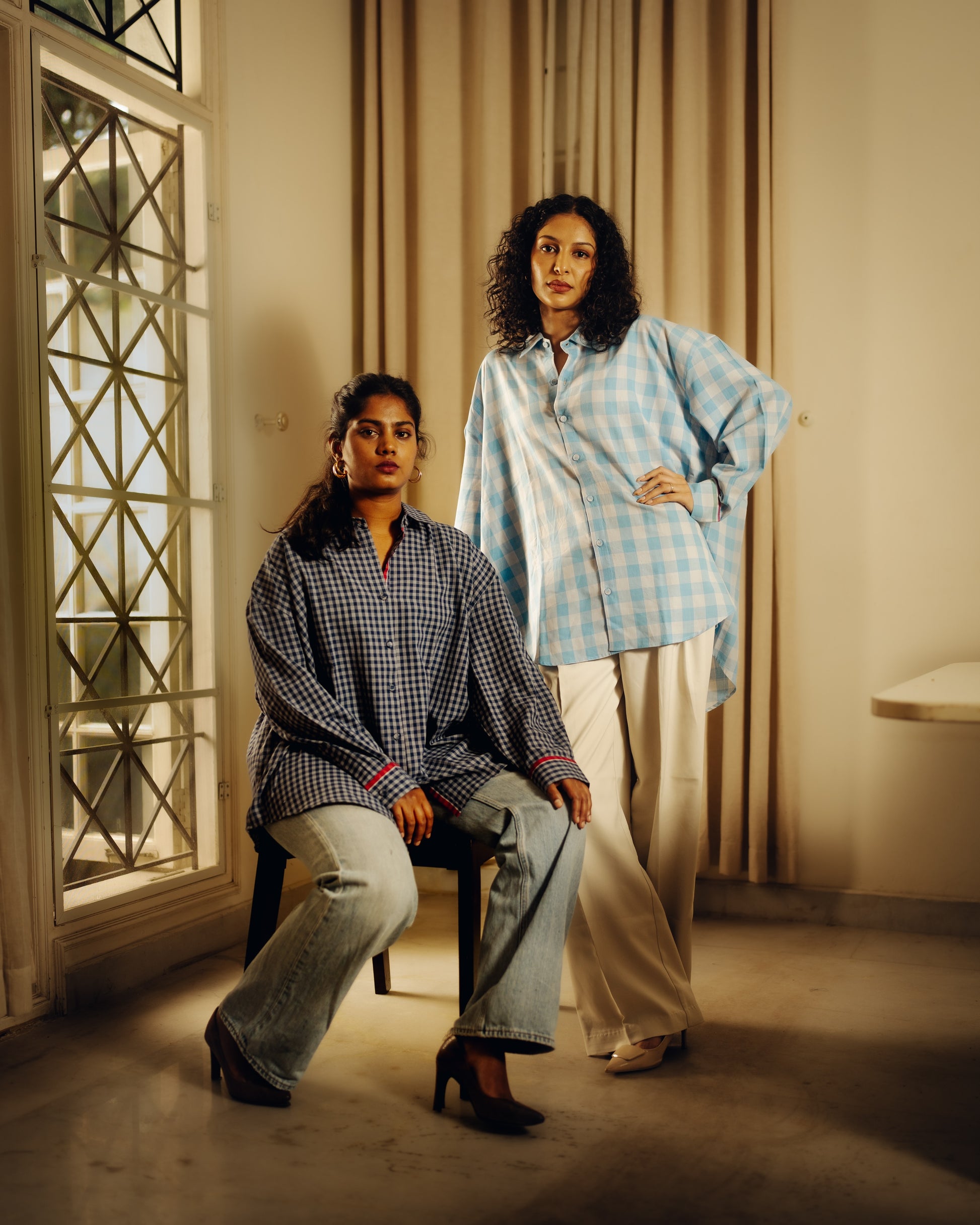 Two women wearing checkered shirts in a room with curtains and a mirror.