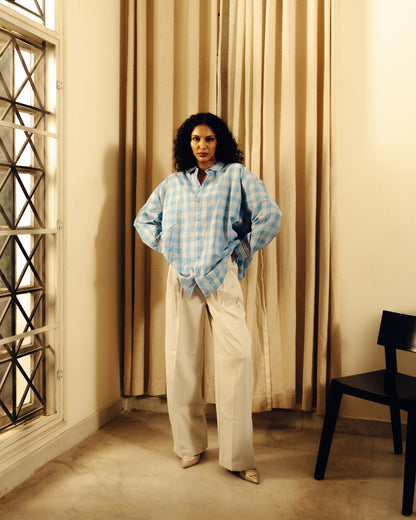 Woman wearing a Oversized shirt - Light blue checks standing in a room with beige curtains and a window.