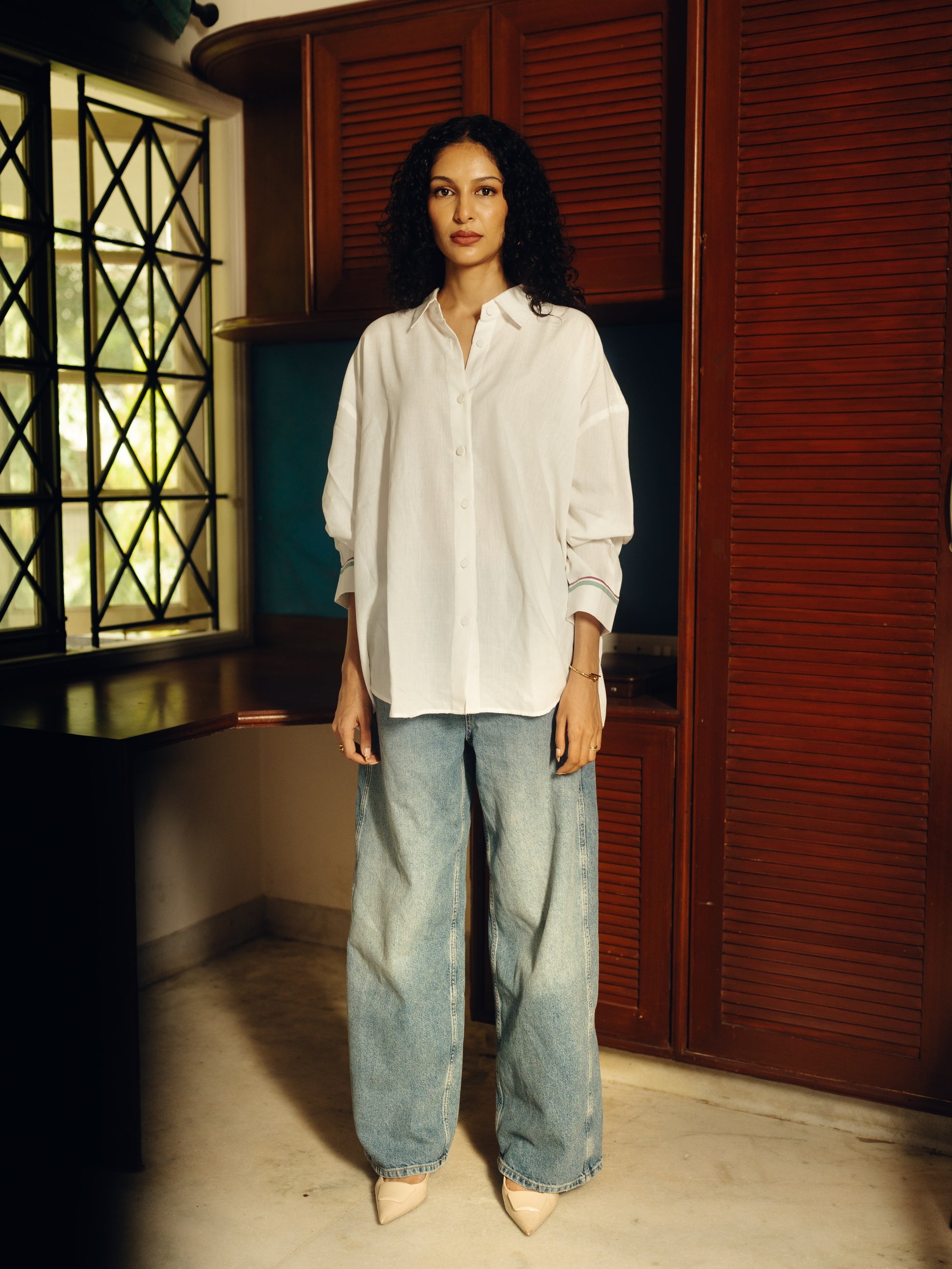Woman wearing a Oversized shirt - White and blue jeans standing in a room with wooden shutters.