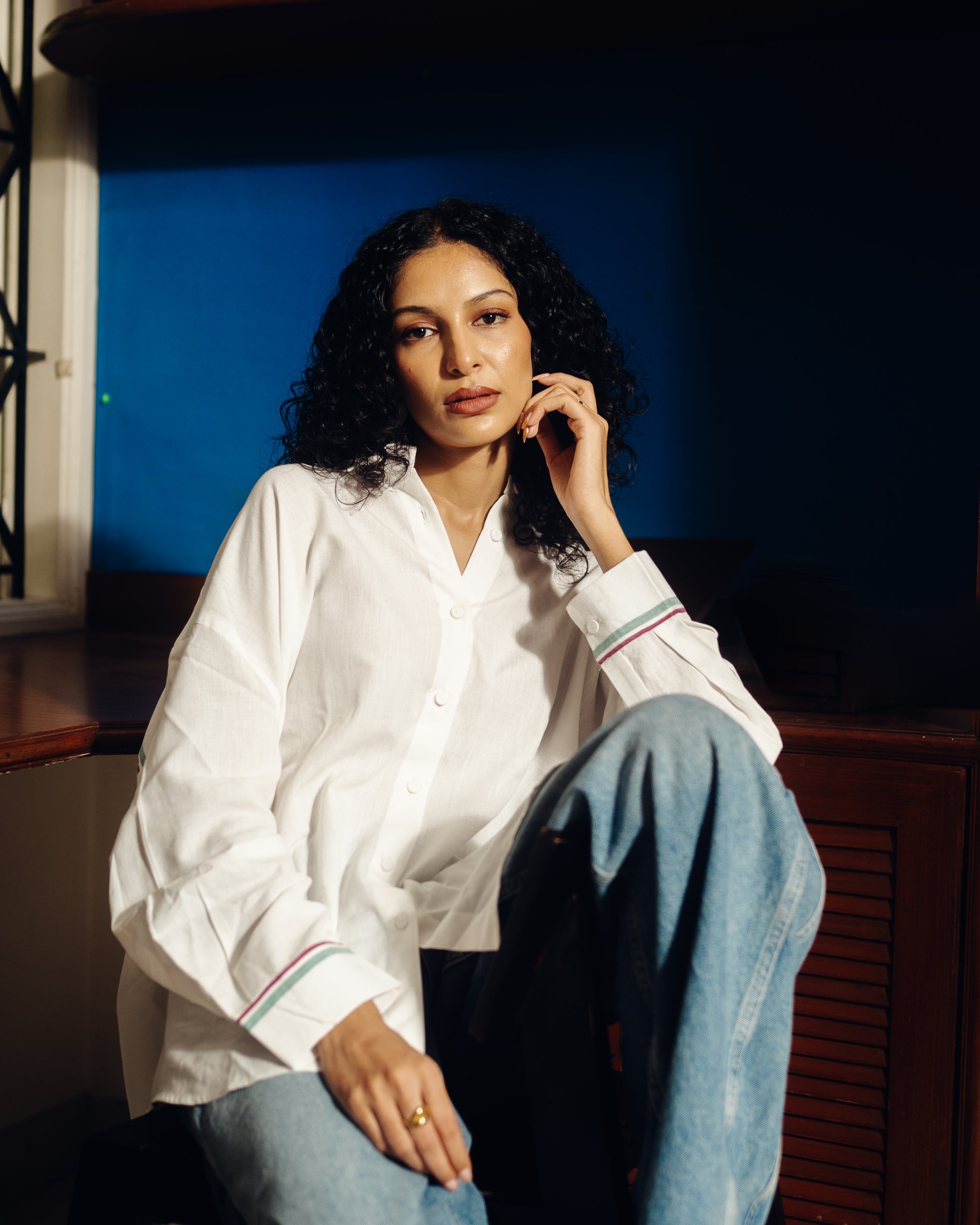 Woman wearing a Oversized shirt - White and blue jeans sitting against a dark background