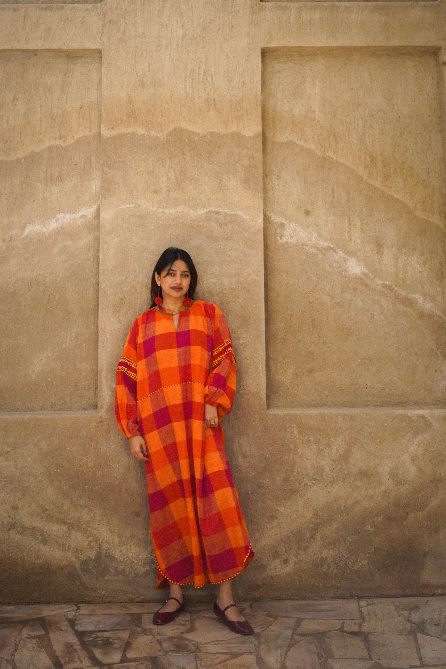 Woman in an orange checkered dress standing against a beige stone wall.