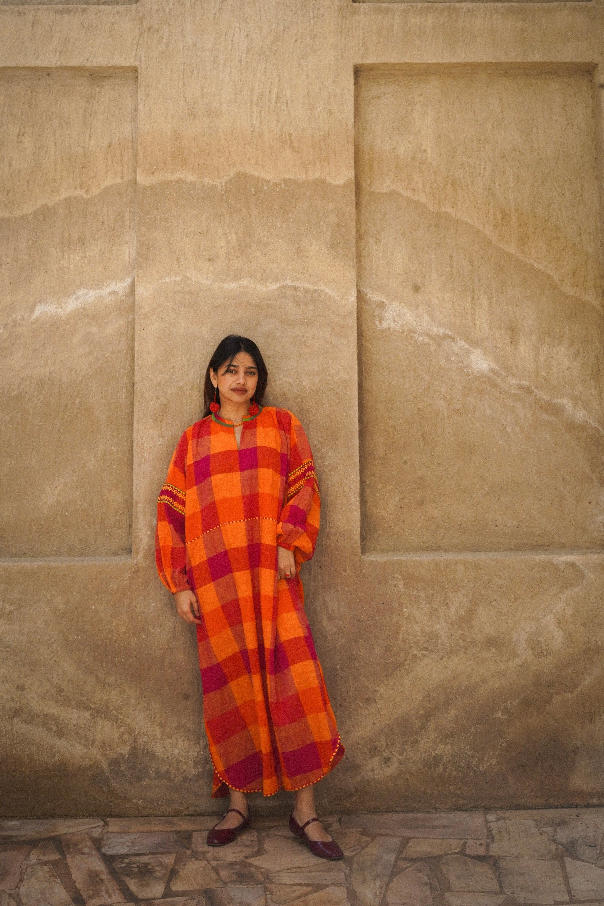 Woman in an orange checkered dress standing against a beige stone wall.