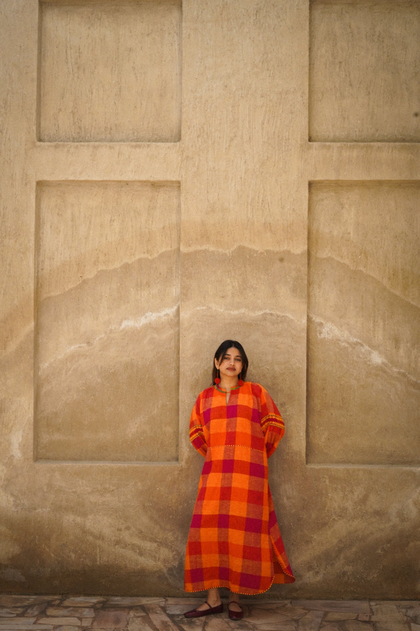 Woman in a red and orange checkered dress standing against a beige stone wall.