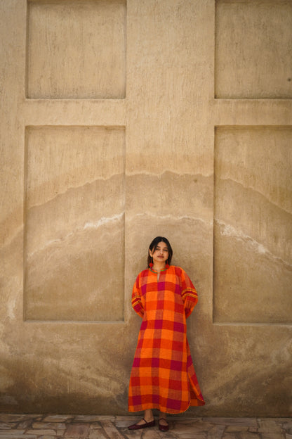 Woman in a red and orange checkered dress standing against a beige stone wall.
