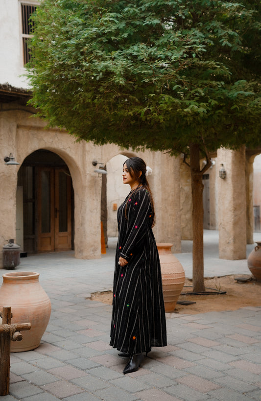 Woman in a long black dress standing in a courtyard with trees and stone walls.