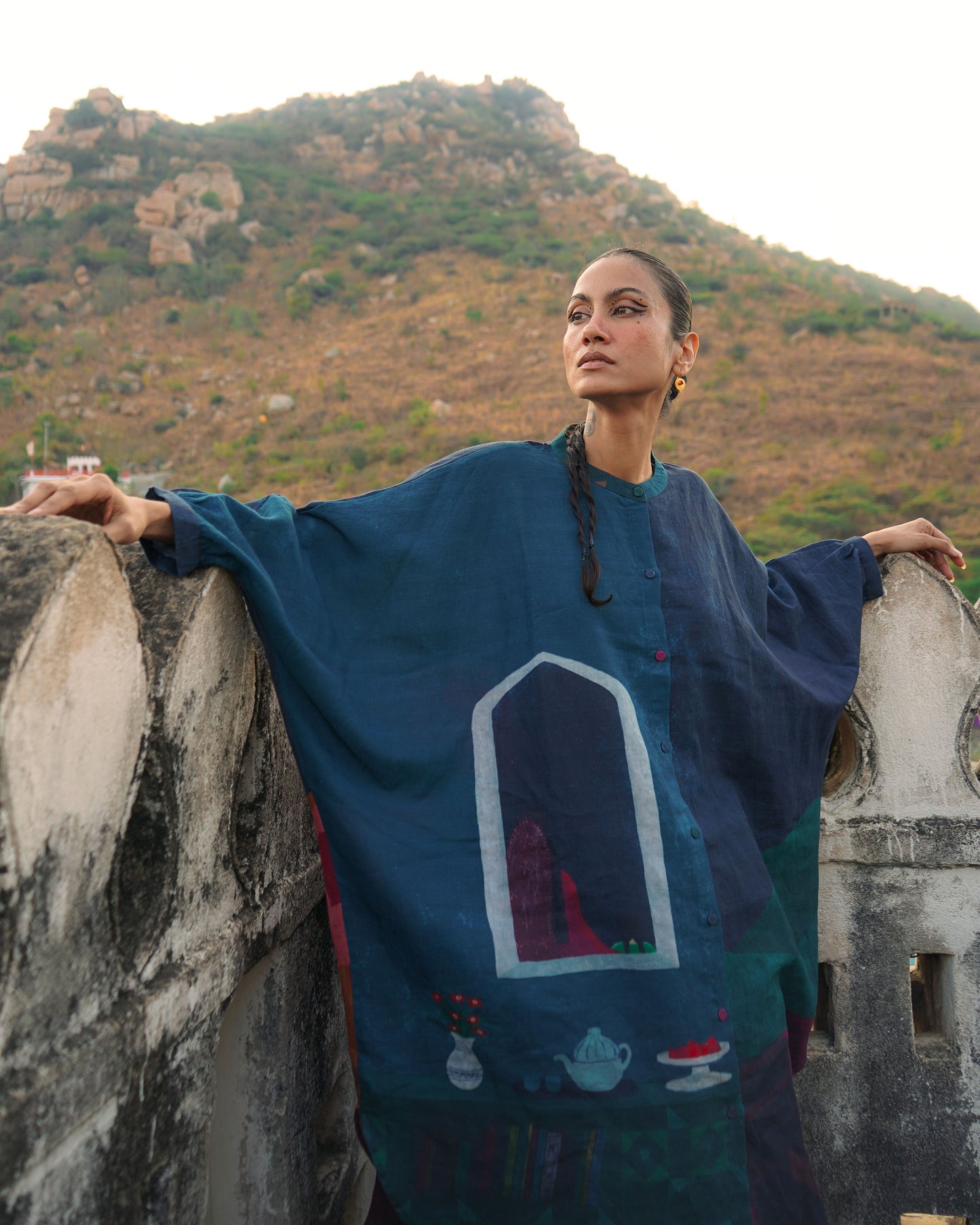 Woman in a blue embroidered dress standing on a stone wall with mountains in the background